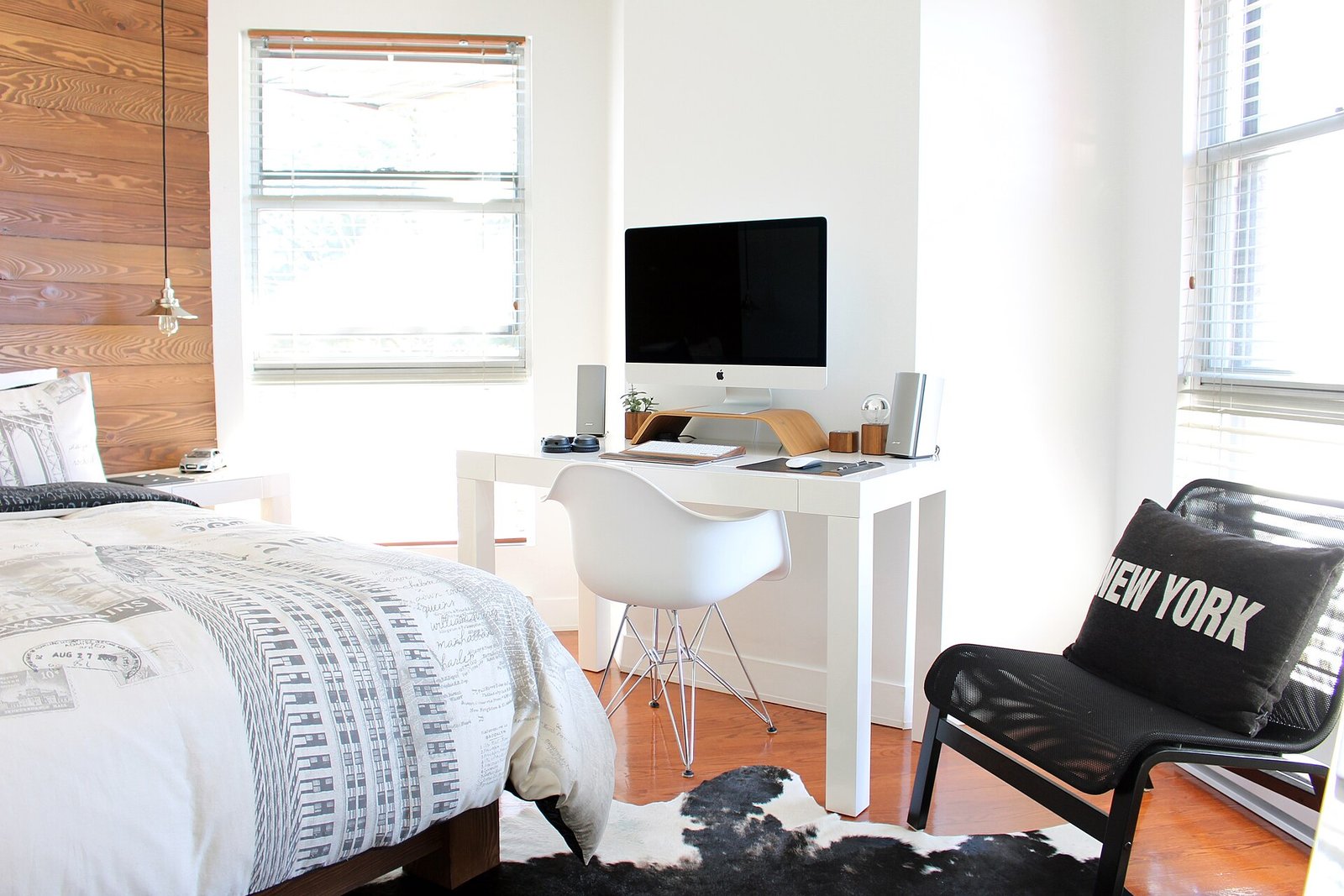 A white bedroom with a patterned duvet, a pine plank wall, a white desk with an iMac and speakers, a white shell chair, a black armchair and a cowhide rug.